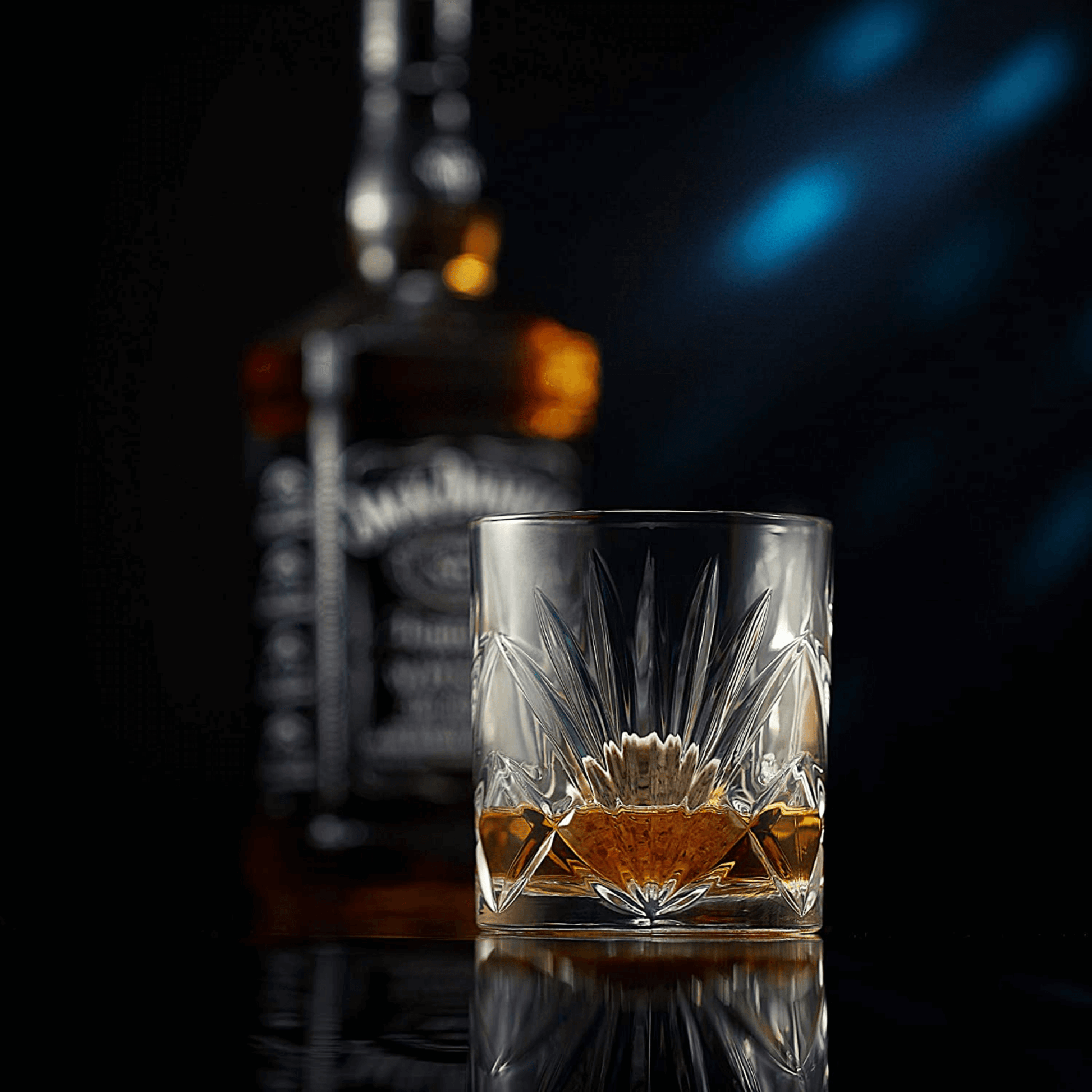 A cut crystal glass from The Connoisseur's Set - Palm Whiskey Glass Edition, filled with whiskey, sits in focus on a reflective surface, with a blurred bottle of Jack Daniels in the background against a dark backdrop.