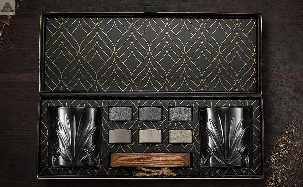 The Connoisseur's Set - Palm Whiskey Glass Edition includes two crystal glasses, six whiskey stones, and a wooden tong, beautifully arranged in a black and gold patterned box on a dark wooden surface.