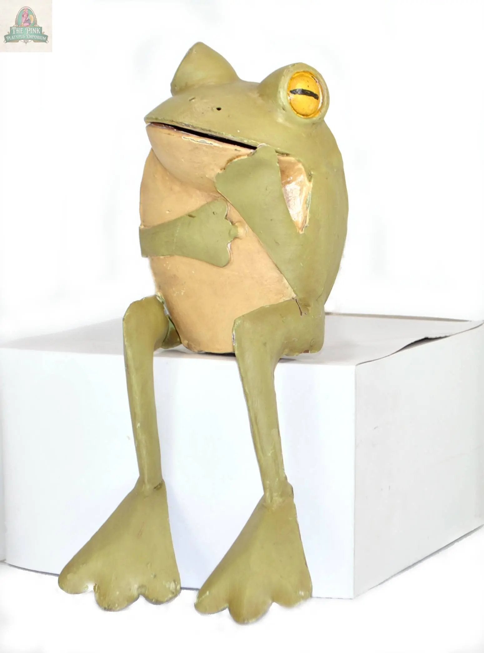 The Thinking Frog figurine, an 11" green and beige hand-painted metal piece with exaggerated features, sits on the edge of a white box, one hand on its chest and the other at its side, against a plain white background.