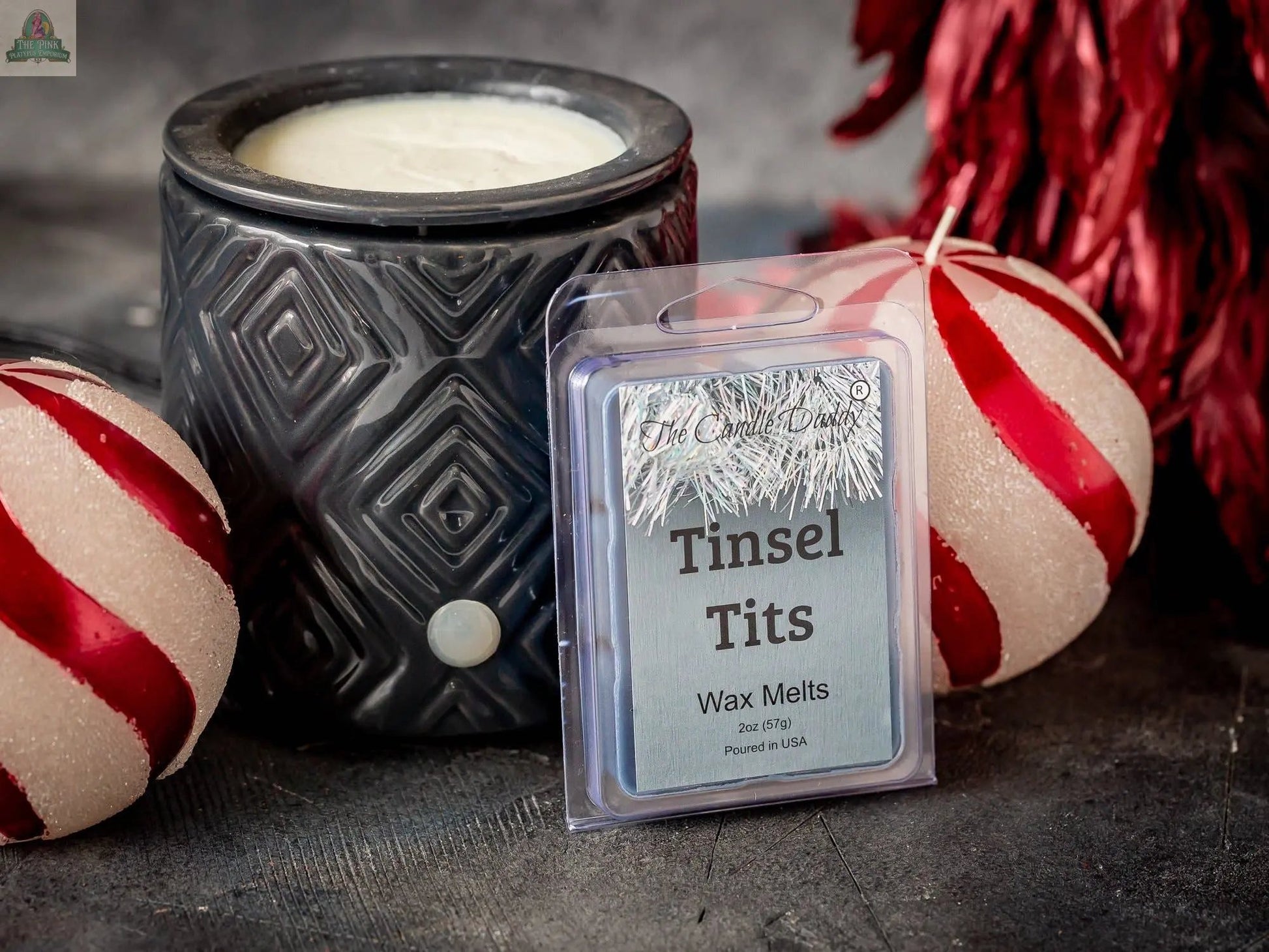 A pack of Tinsel Tits - Mountain Top Tease Scented Wax Melts is shown in front of a black candle warmer, with two red and white striped Christmas ornaments and festive red foliage in the background.