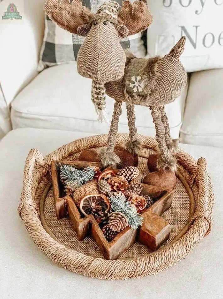 A fabric moose figurine stands on the Handmade Tree Dough Bowl (#A), filled with pinecones, dried orange slices, and evergreen branches, and displayed on a woven tray atop a beige couch.