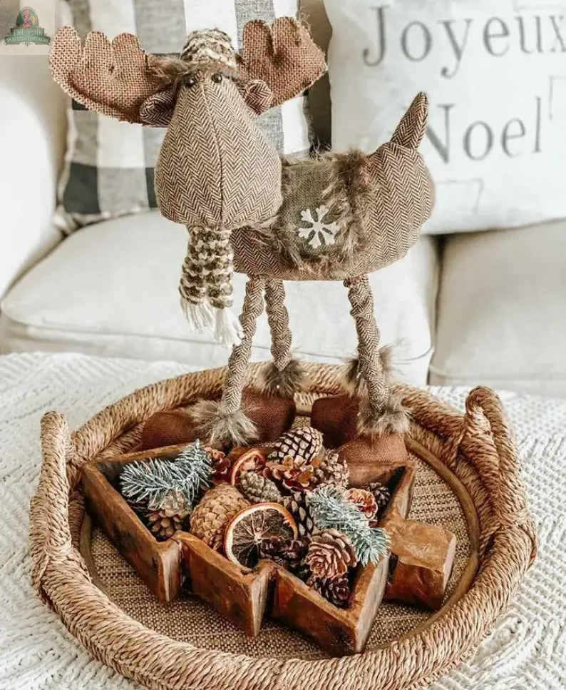 A plush reindeer stands on a woven tray filled with pinecones, dried citrus, and fir in a handmade Tree Dough Bowl (#A), star-shaped, with festive pillows in the background.