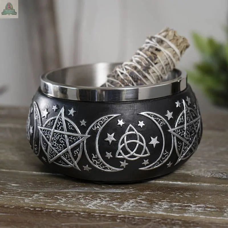 The Triple Moon Resin Smudge Ritual Offering Bowl, black with silver pentagrams, moons, and stars, elegantly holds a sage bundle tied with string—ideal for smudging rituals and displayed beautifully on a wooden surface.