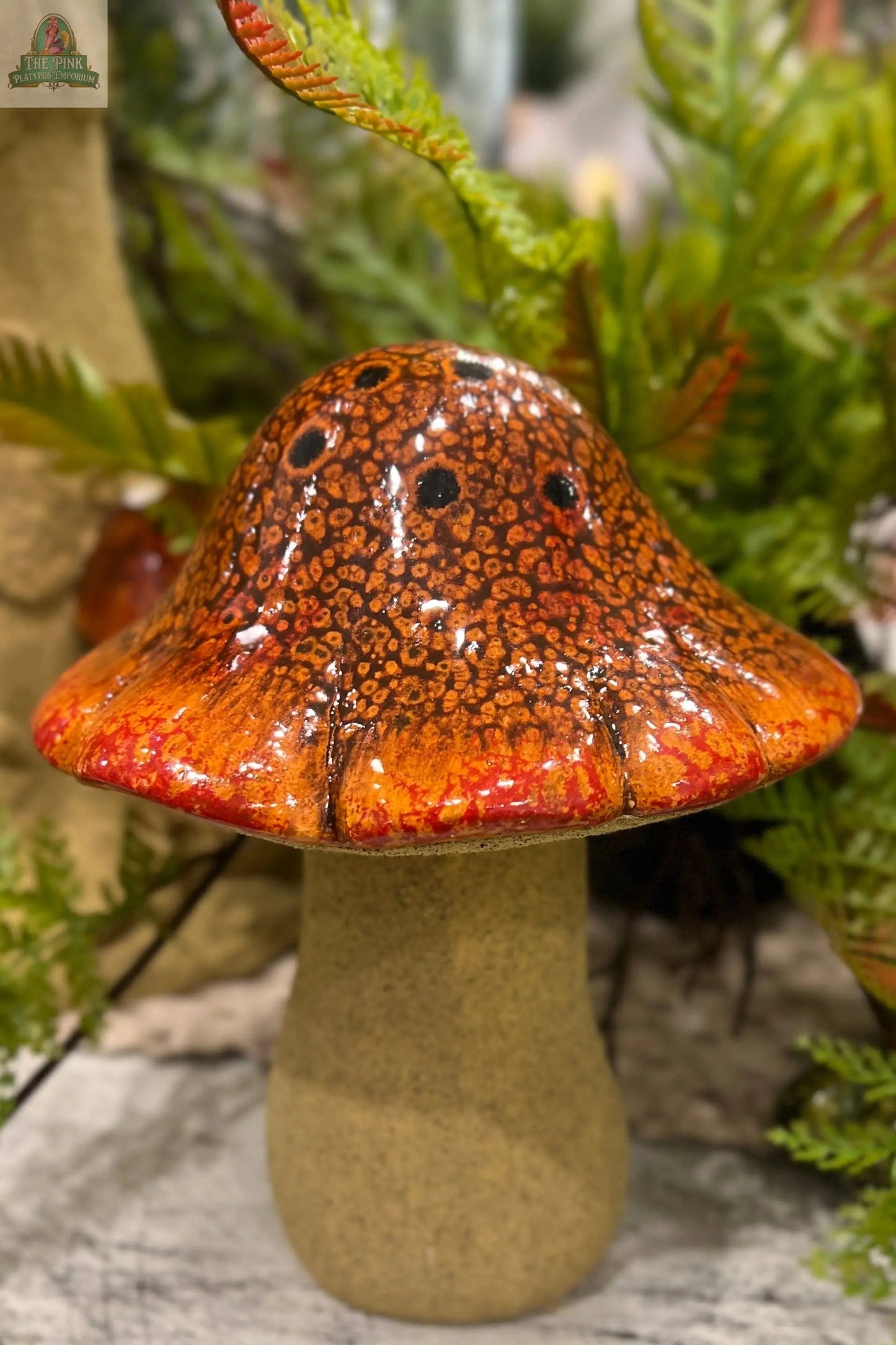 The Umbrella Mushroom sculpture (5x5x6.4in) features a textured orange and brown ceramic cap, a beige stem, and is accented by green ferns and foliage.