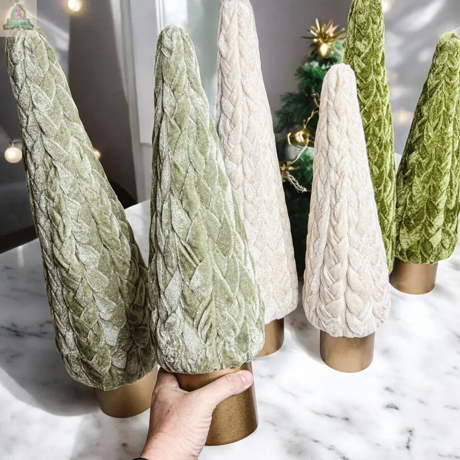 A hand holds a Velvet Christmas Tree in soft, textured light green velvet, displayed beside similar plush trees in light green and cream with gold bases—ideal holiday decor for a farmhouse-style display on a white marble surface.