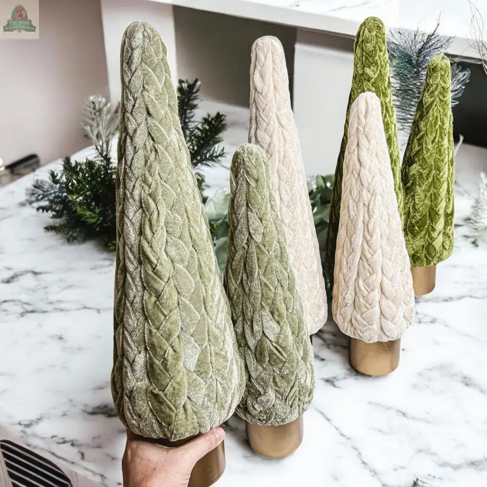Six Velvet Christmas Trees in green and cream are arranged on a marble countertop with greenery in the background. A hand holds one of the green Velvet Christmas Tree decorations in the foreground, ideal for cozy holiday decor.