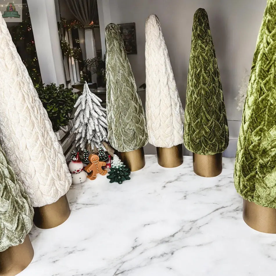A marble table showcases Velvet Christmas Trees in green and cream with gold bases. Festive figurines, a white tree, and sparkling holiday décor add a farmhouse touch in the background.