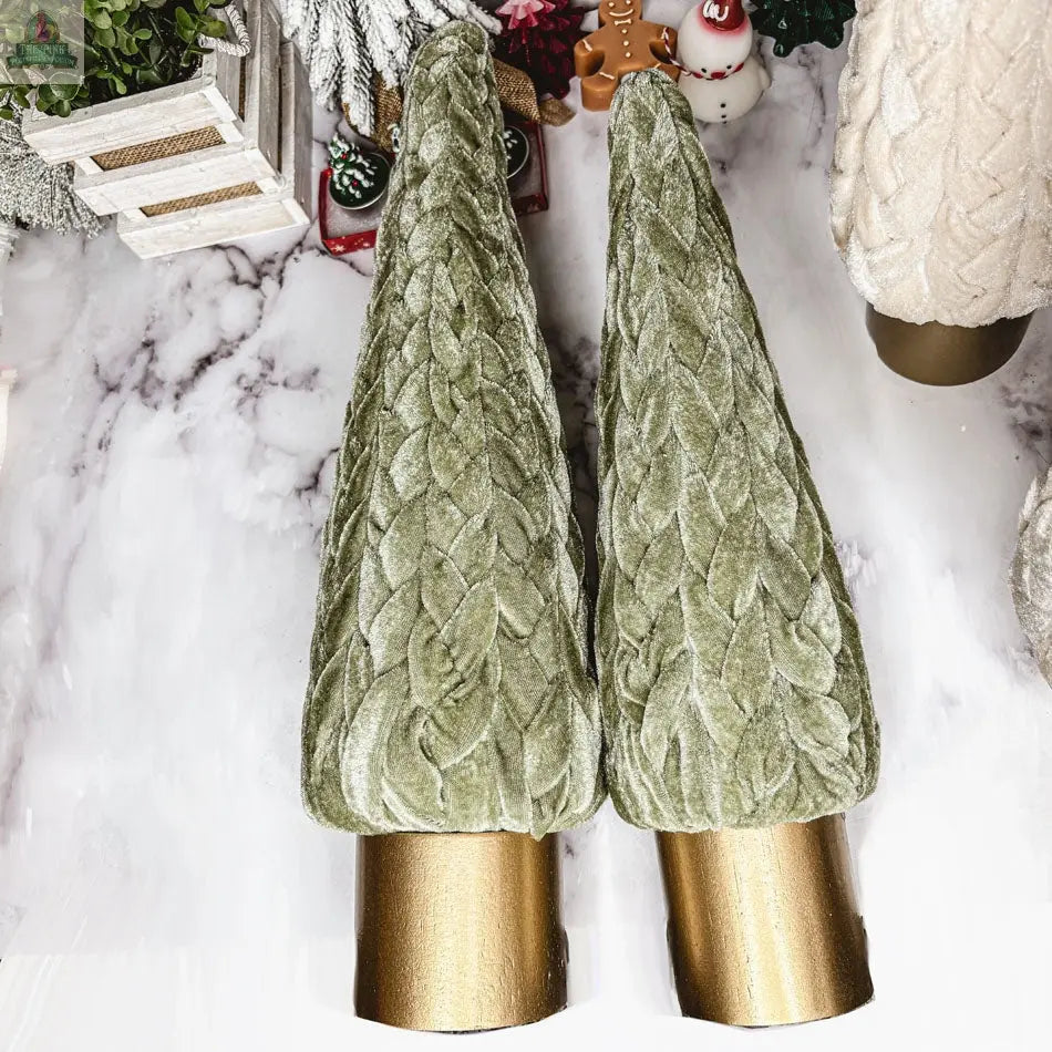 Two Velvet Christmas Trees in green, featuring a textured leaf pattern and gold cylindrical bases, stand upright on a white marble surface amid other holiday décor.