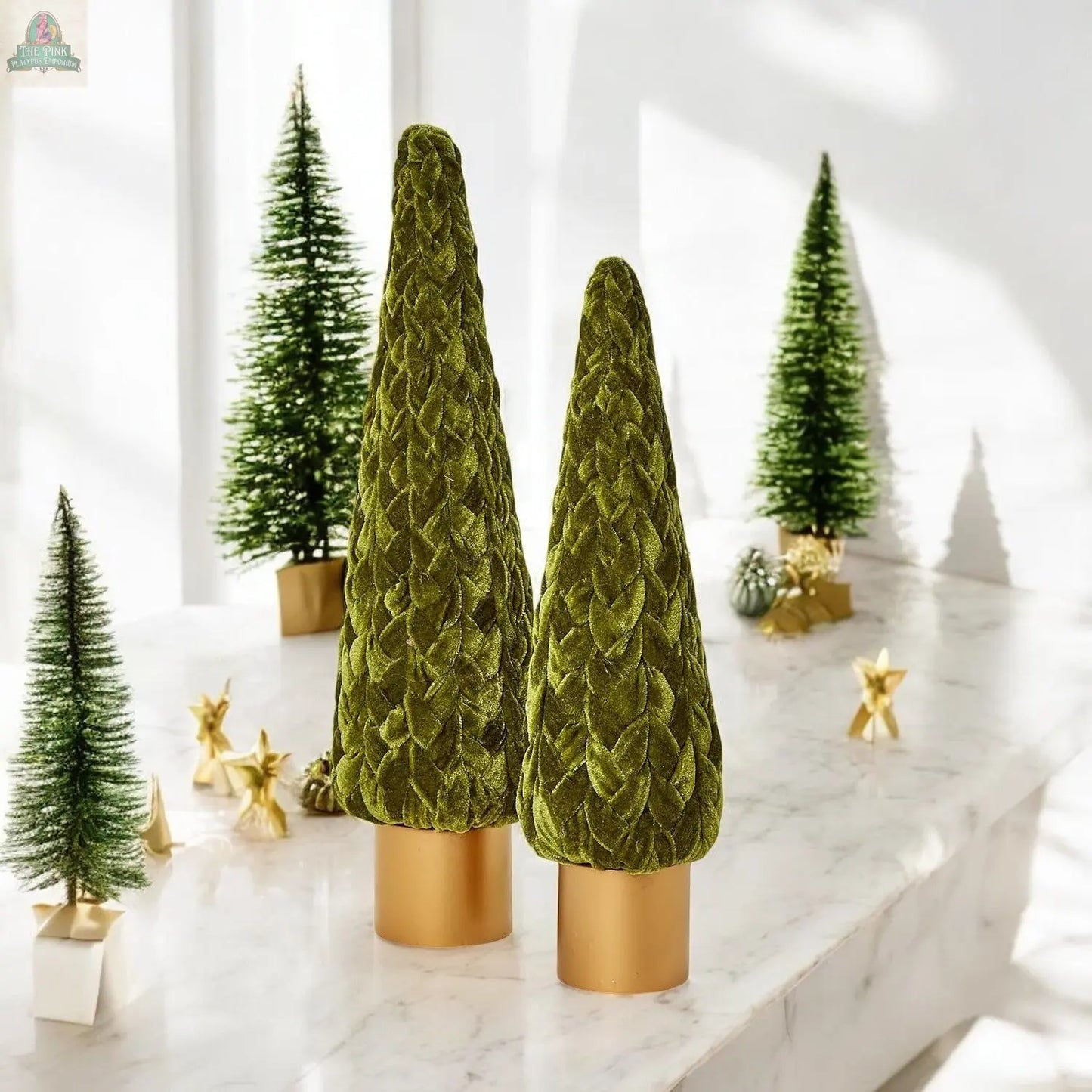 Two Velvet Christmas Trees with gold bases stand on a marble surface, surrounded by holiday décor like artificial pine trees and gold star ornaments, creating a festive mantel display in a brightly lit room.