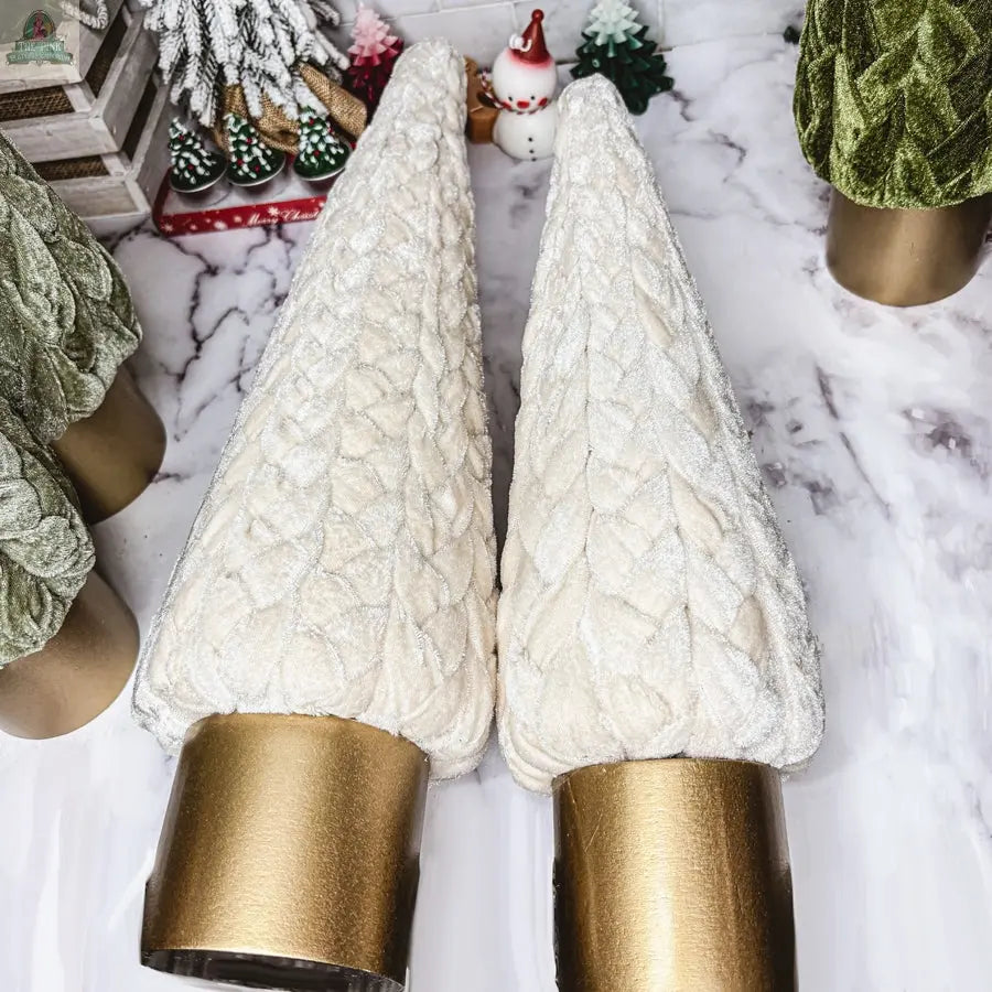 Two cream-colored Velvet Christmas Trees with gold bases sit on a marble surface. Green decorations, a Santa figurine, and festive ornaments complete the holiday décor.