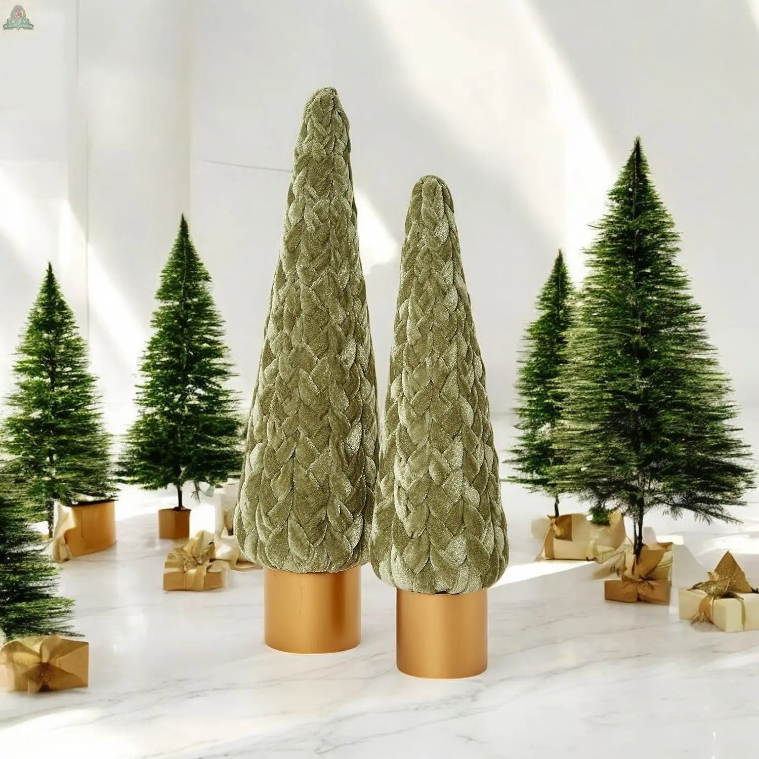 Two Velvet Christmas Trees with textured green velvet surfaces on gold bases are displayed among festive décor, including small faux pine trees and gold-wrapped gifts, all arranged on a white floor in a bright room.