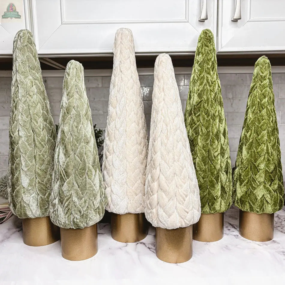 Six Velvet Christmas Trees with textured leaf-like patterns in green and cream stand on a countertop, each with a gold cylindrical base, creating charming holiday décor against white cabinets and a tiled backsplash.