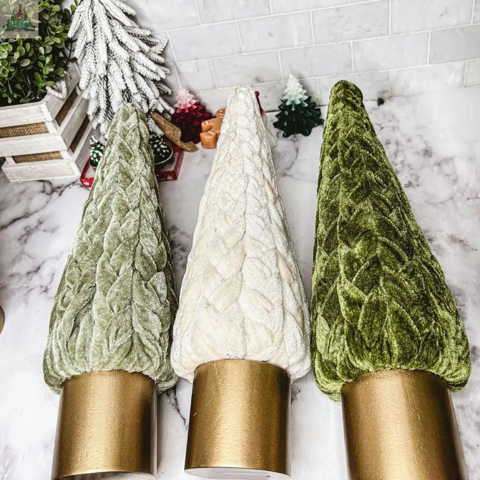 Three Velvet Christmas Trees in green and white shades, each with a gold cylindrical base, are arranged on a marble surface surrounded by miniature trees and festive décor.