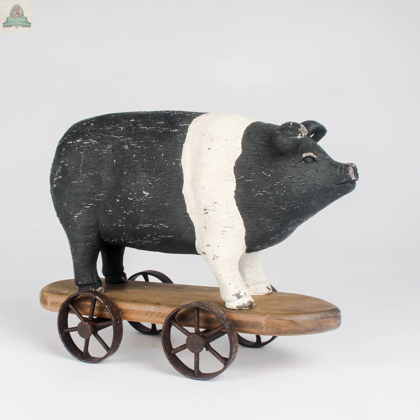 The Vintage Farmhouse Pig on Wheels by Pink Platypus Emporium features a black and white pig figurine with a white band, set on a wooden platform with four metal wheels—a charming farmhouse décor pull toy.