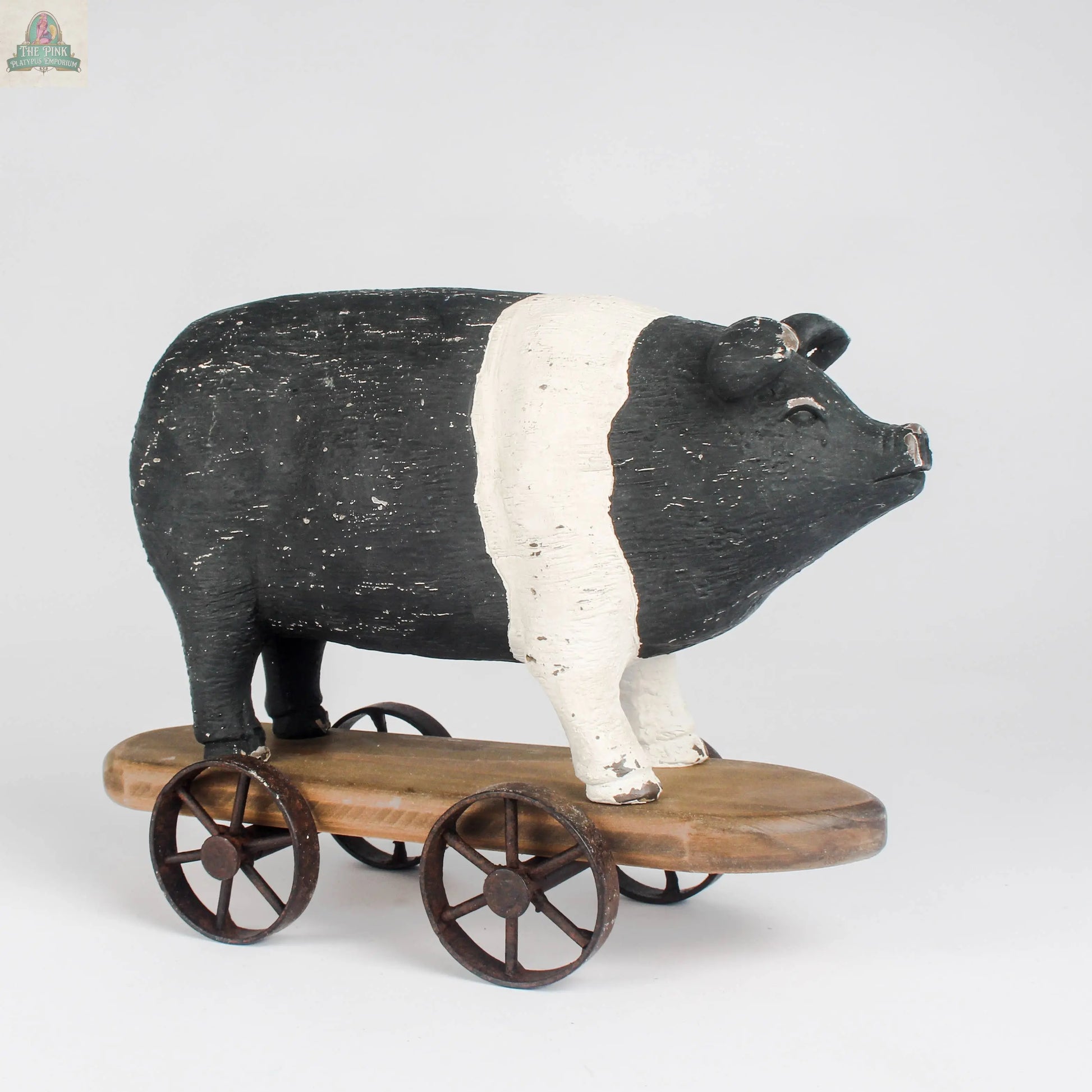 The Vintage Farmhouse Pig on Wheels by Pink Platypus Emporium features a black and white pig figurine with a white band, set on a wooden platform with four metal wheels—a charming farmhouse décor pull toy.