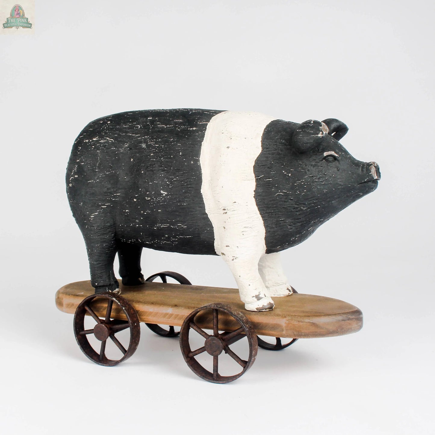 The Pink Platypus Emporium Vintage Farmhouse Pig on Wheels is a black and white pig figurine on a wooden platform with four metal wheels, evoking a classic pull toy, shown against a plain white background.