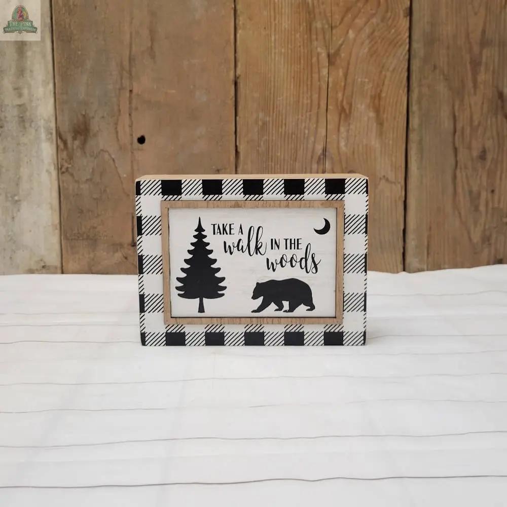 The Walk in the Woods sign is a framed hanging decor with a black-and-white plaid border, silhouettes of a tree, bear, and crescent moon, and the phrase “Take a walk in the woods,” perfect for rustic table top displays.