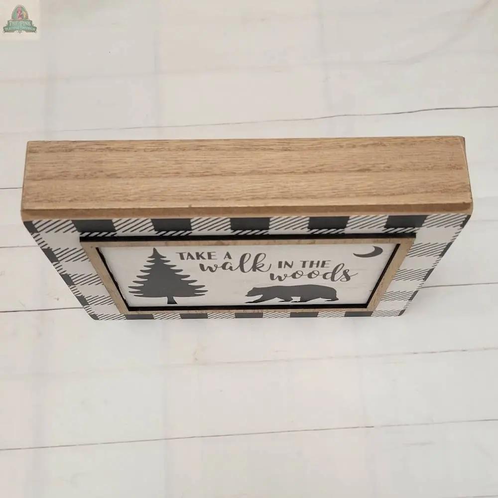 The Walk in the Woods sign is a decorative wooden hanging piece with a checkered border, featuring a tree, bear, and crescent moon, and the phrase "Take a walk in the woods"—ideal for charming home decor on any light-colored tabletop.