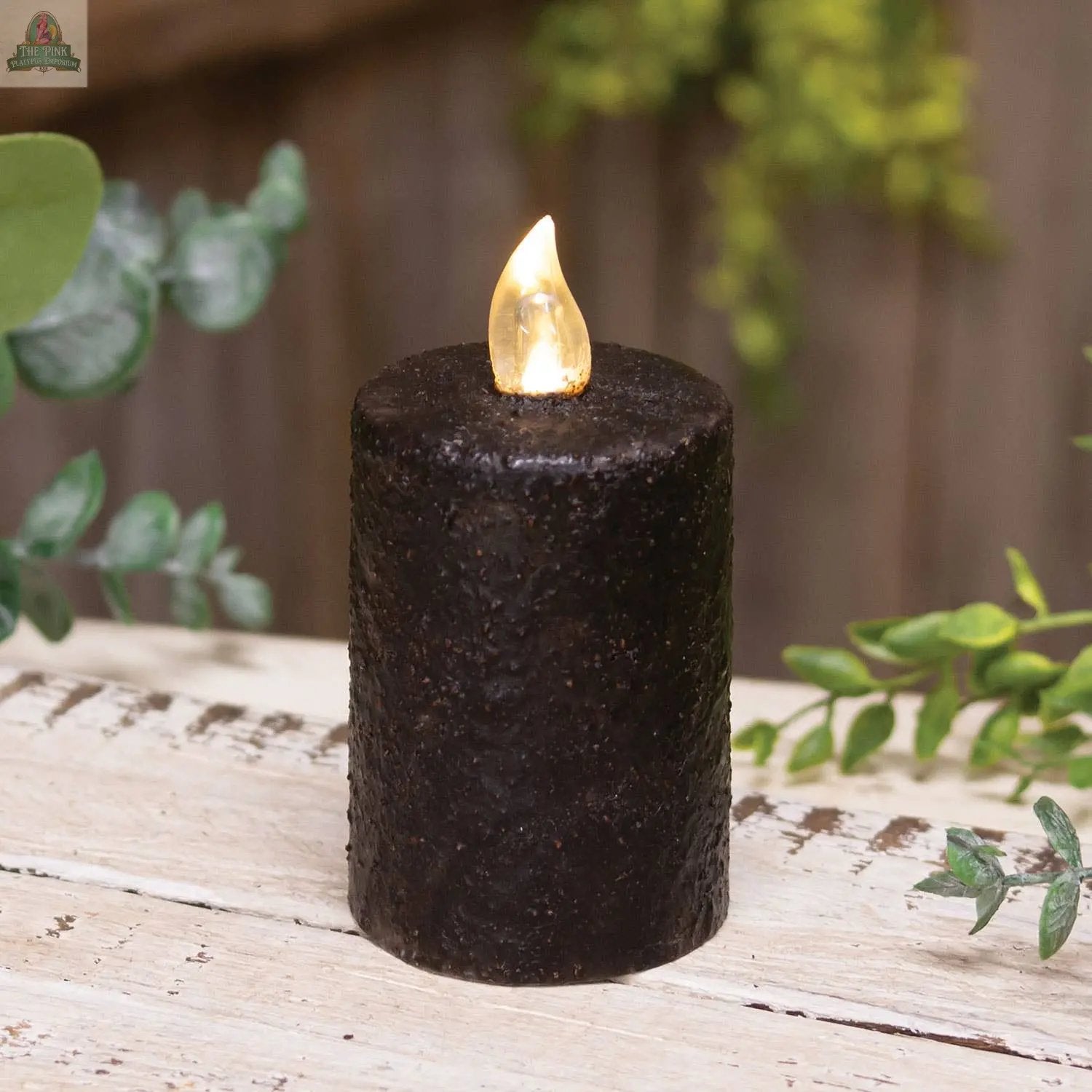 The Warm Glow Textured Black Flicker Flame Timer Pillar, 3", sits on a rustic white wooden surface among green leaves and blurred plants, creating a cozy, elegant ambiance.