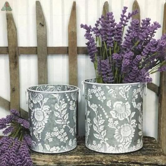Two White Embossed Flower Containers with white floral patterns sit on a wooden surface; one holds purple lavender flowers, and a rustic wooden fence stands in the background.