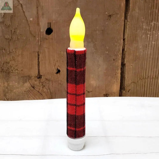 The White Timered LED Taper with Red Buffalo Check Wrap stands upright on a white surface, its yellow flame glowing against a rustic wooden background.