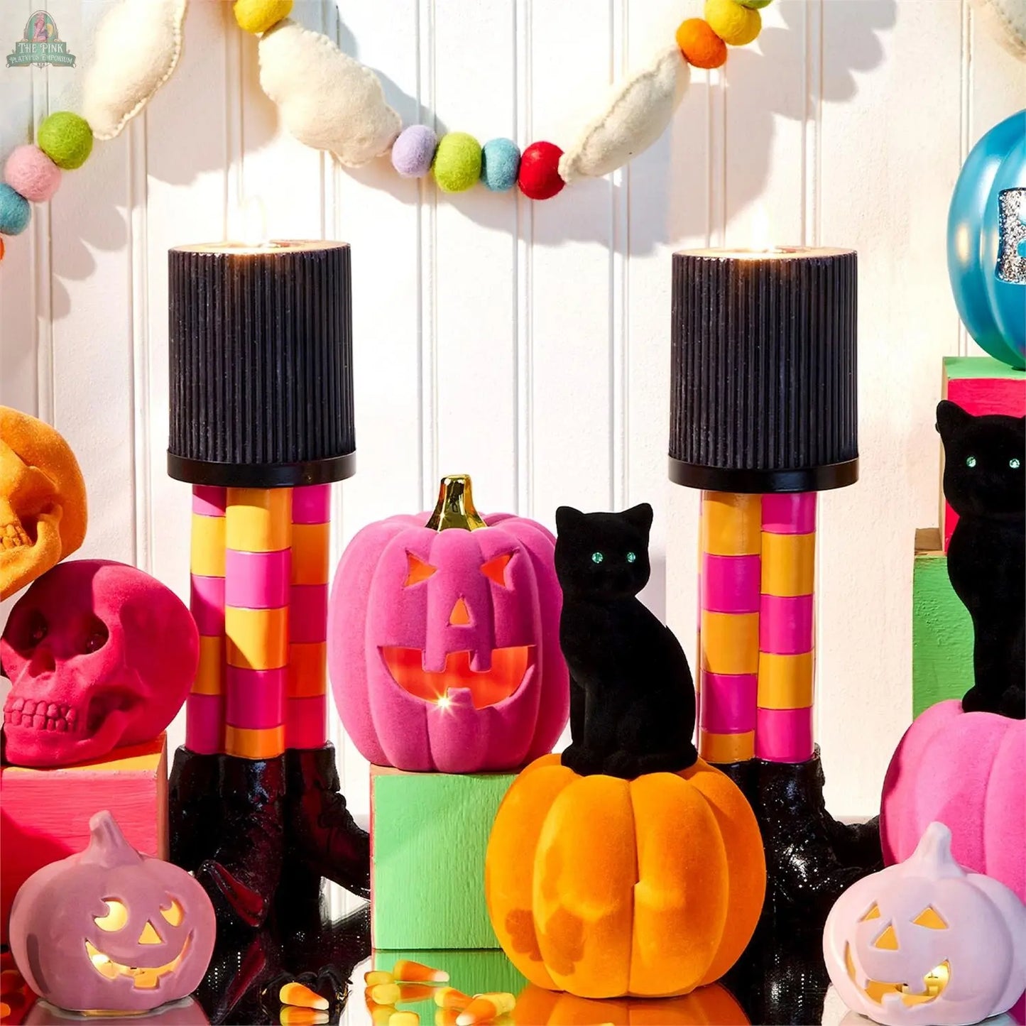 Colorful Halloween decor with velvet pumpkins, black cat, pink skull, Wicked Witch Candle Holder with striped legs and black tops, garland, and candy corn arranged against a white wood-paneled background.