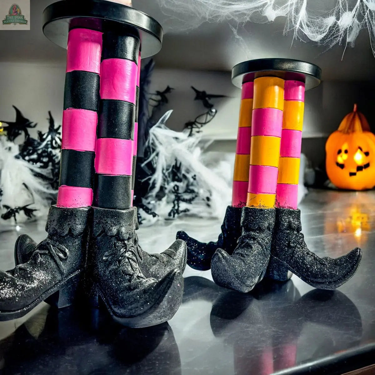 Two Wicked Witch Candle Holders, one with pink and black striped legs, the other with yellow and pink, make a whimsical Halloween centerpiece. Pair them with festive decor like a glowing jack-o’-lantern for a perfectly spooky scene.