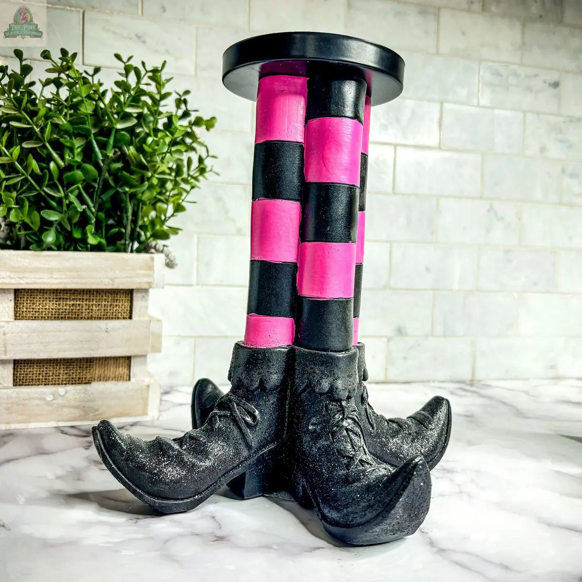 The Wicked Witch Candle Holder is a decorative Halloween centerpiece shaped like two legs in black and pink stripes with curled shoes, perfect for tabletops.