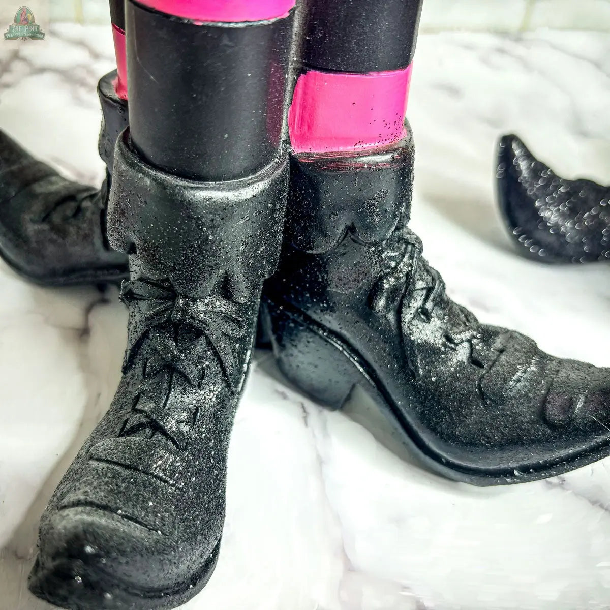 Close-up of the Wicked Witch Candle Holder: black, shiny, lace-up boots with pointed toes over black pants with a pink stripe, standing on a white textured surface—an eye-catching piece for unique Halloween decor.