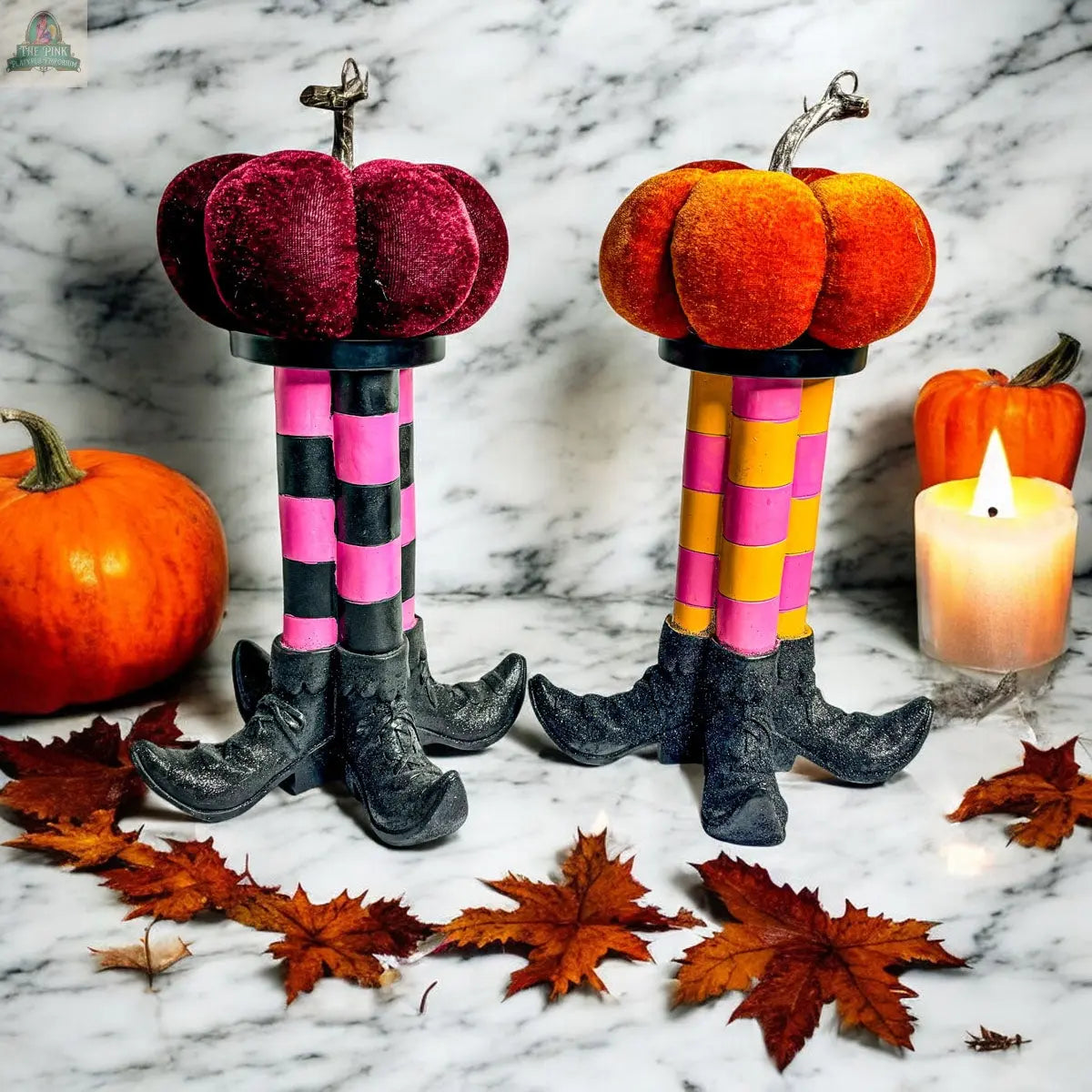 Two Wicked Witch Candle Holders—whimsical centerpieces shaped like witch legs with velvet pumpkins on top, one pink/black and one yellow/purple—are displayed among pumpkins, autumn leaves, and a lit candle on a marble surface.