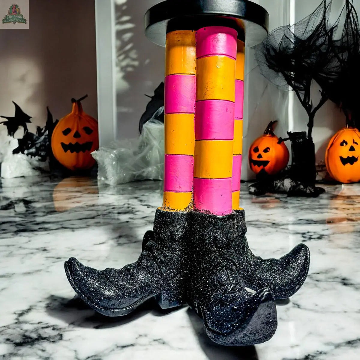 The Wicked Witch Candle Holder features pink and yellow striped legs with glittery black shoes. Surrounded by jack-o-lanterns and spider webs, this whimsical piece makes a festive Halloween centerpiece.