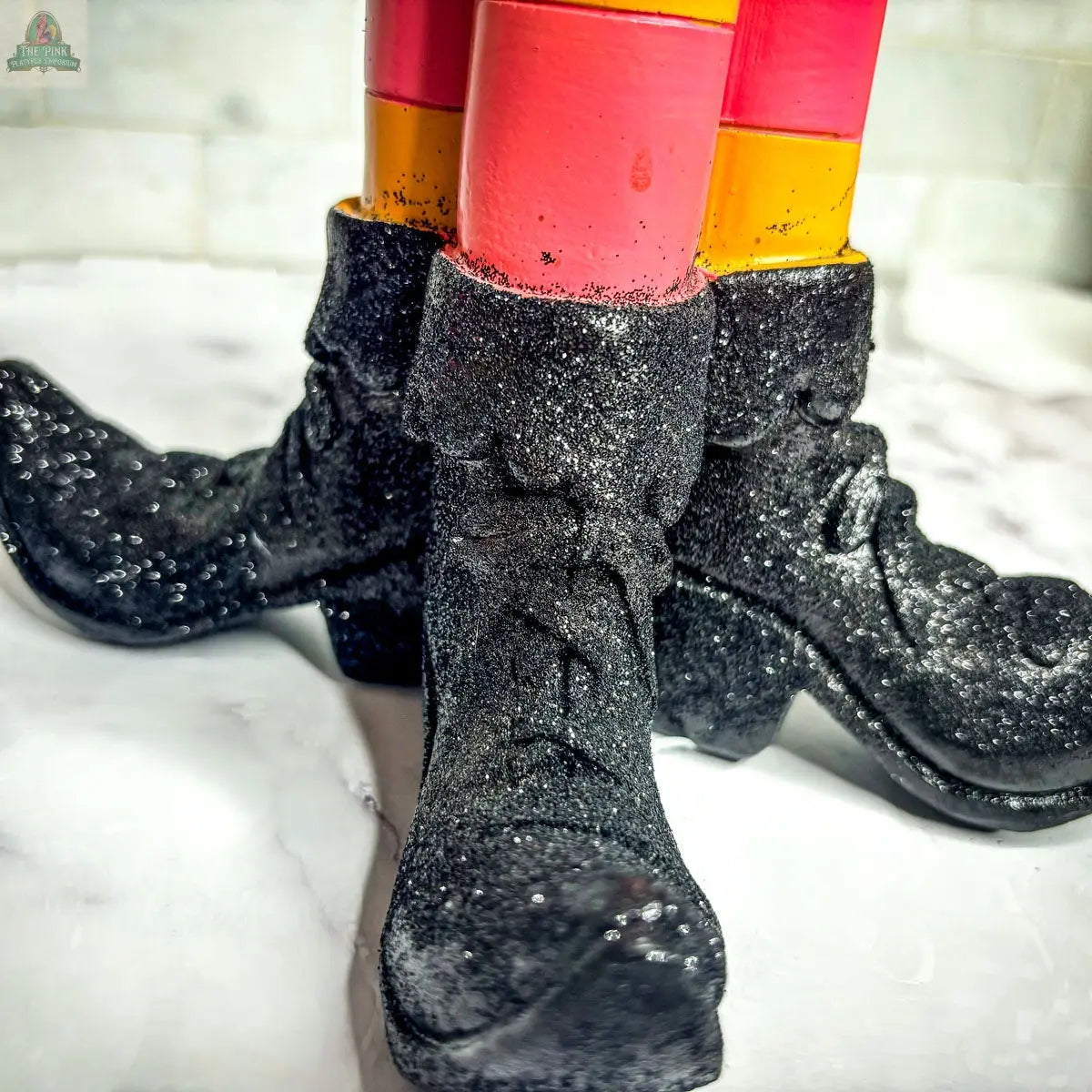 The Wicked Witch Candle Holder features three pairs of black, glittery shoes with pink and yellow striped legs on a white marble base—an eye-catching Halloween centerpiece or quirky decor accent.