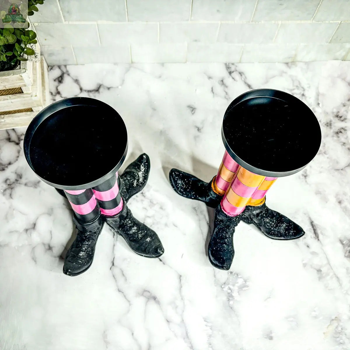 A pair of Wicked Witch Candle Holders, featuring black boot-shaped legs and colorful striped stockings, creates a whimsical Halloween centerpiece on a white marble surface set against a tiled wall.