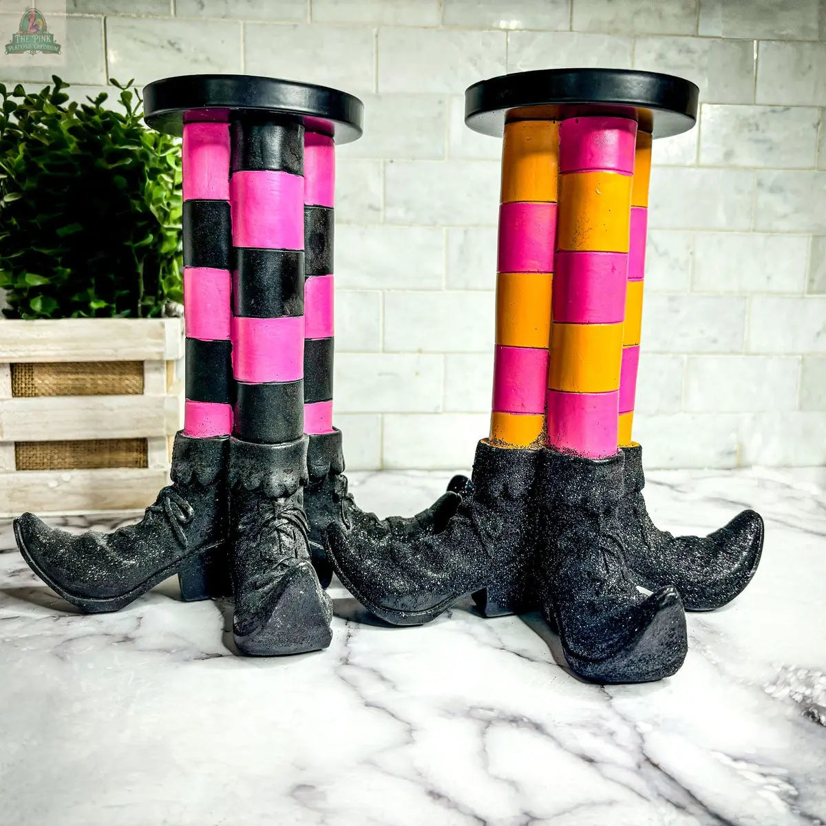Two Wicked Witch Candle Holders—one with pink and black stripes, the other in yellow and orange—stand out on a marble countertop, creating a playful Halloween centerpiece for your spooky decor.