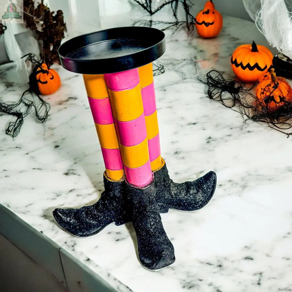 The Wicked Witch Candle Holder features a round tray atop colorful, checkered witch legs and boots—perfect as a Halloween centerpiece on a marble surface surrounded by pumpkins and festive decor.