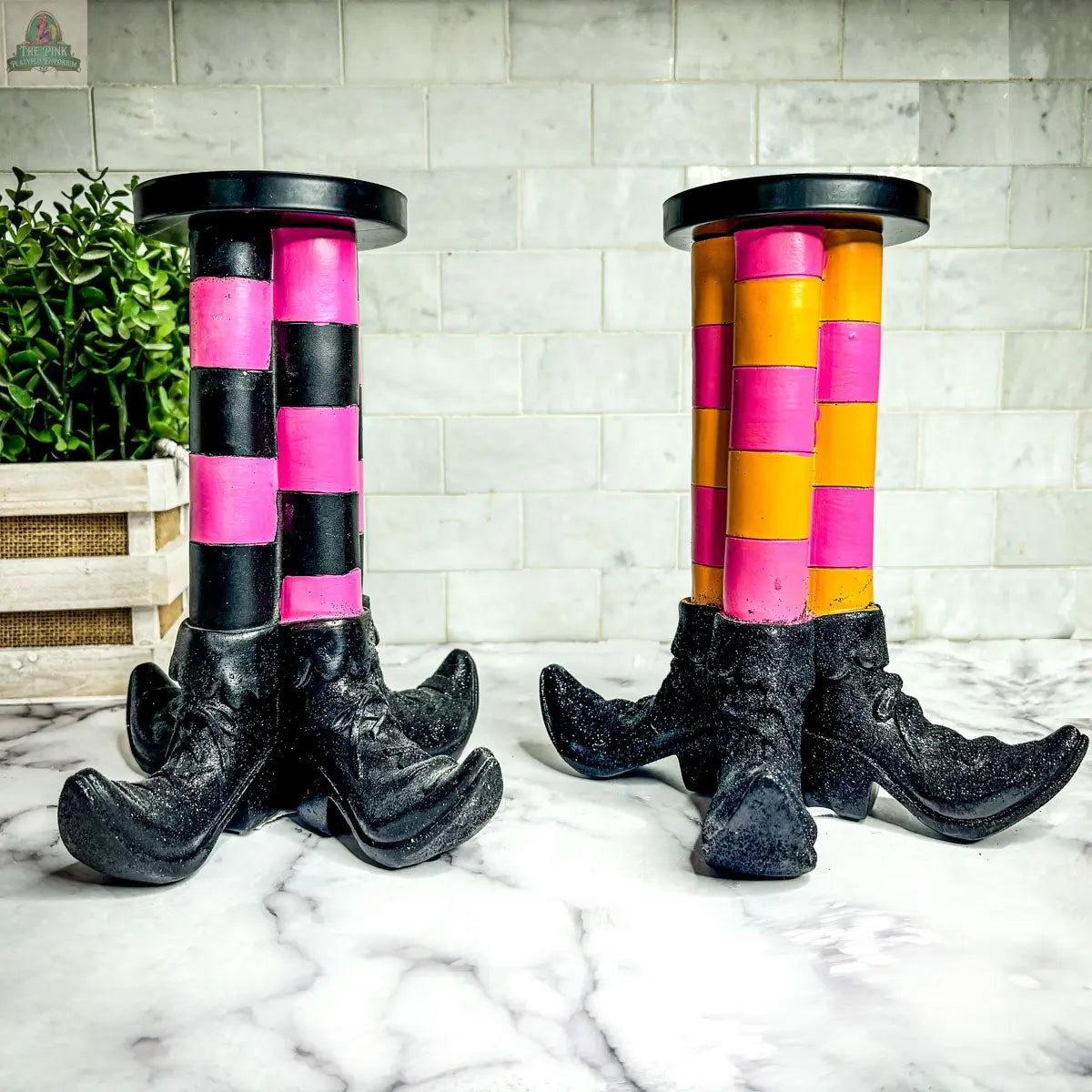 The Wicked Witch Candle Holder, with colorful, striped legs and black witch boot bases, sits on a marble counter with a potted plant and tile backsplash—an ideal touch of playful Halloween decor.