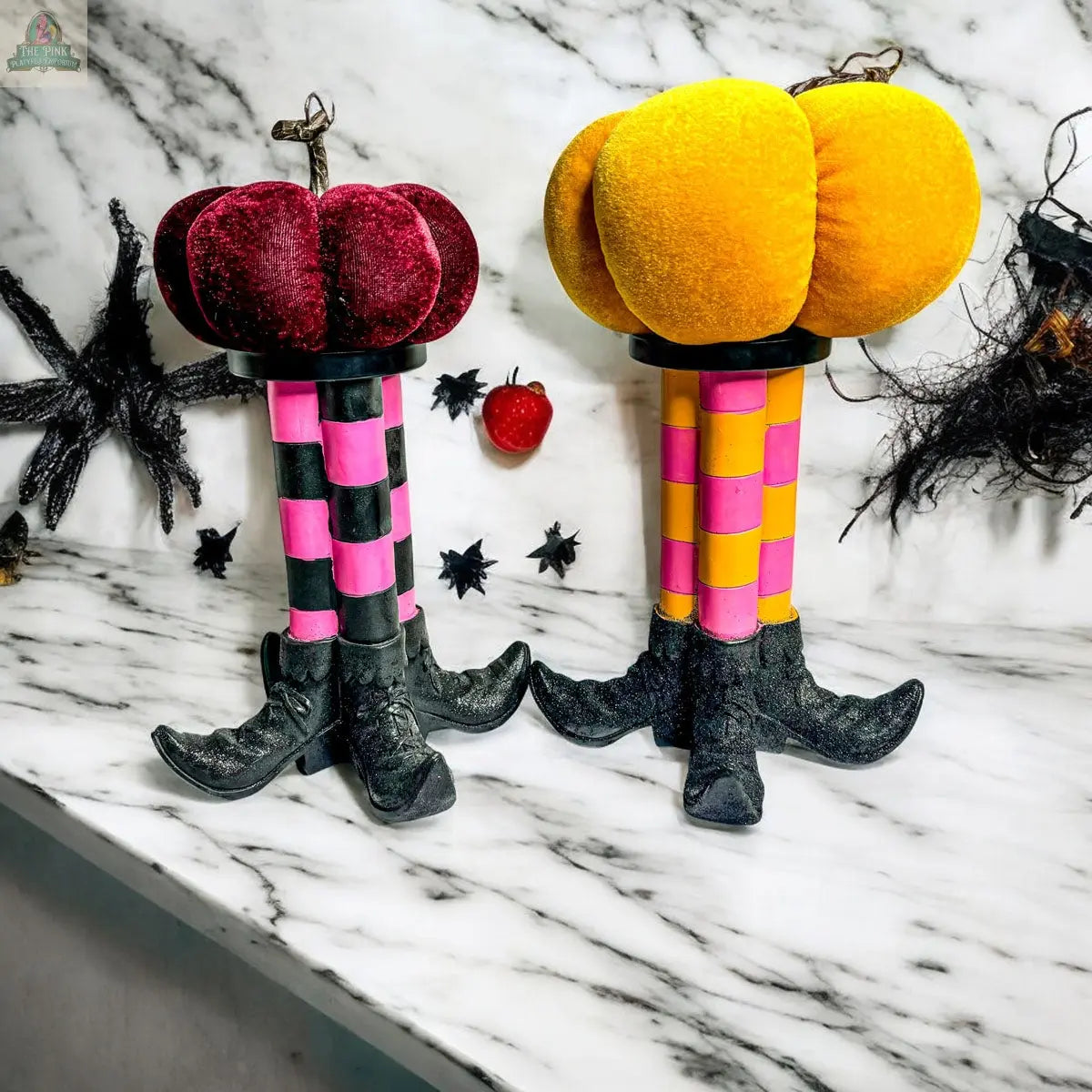 The Wicked Witch Candle Holder features whimsical pumpkin decorations with plush tops on striped pink and yellow stems with black clawed feet and boots, plus scattered spider and bat accents on a marble countertop for a festive Halloween centerpiece.