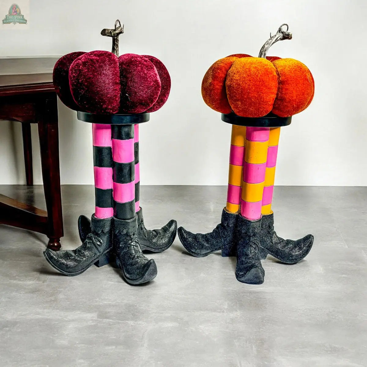 A Wicked Witch Candle Holder adds charming Halloween flair next to a wooden table on a gray floor, complementing whimsical pumpkin-shaped stools with plush seats and checkered legs styled like black witch boots.