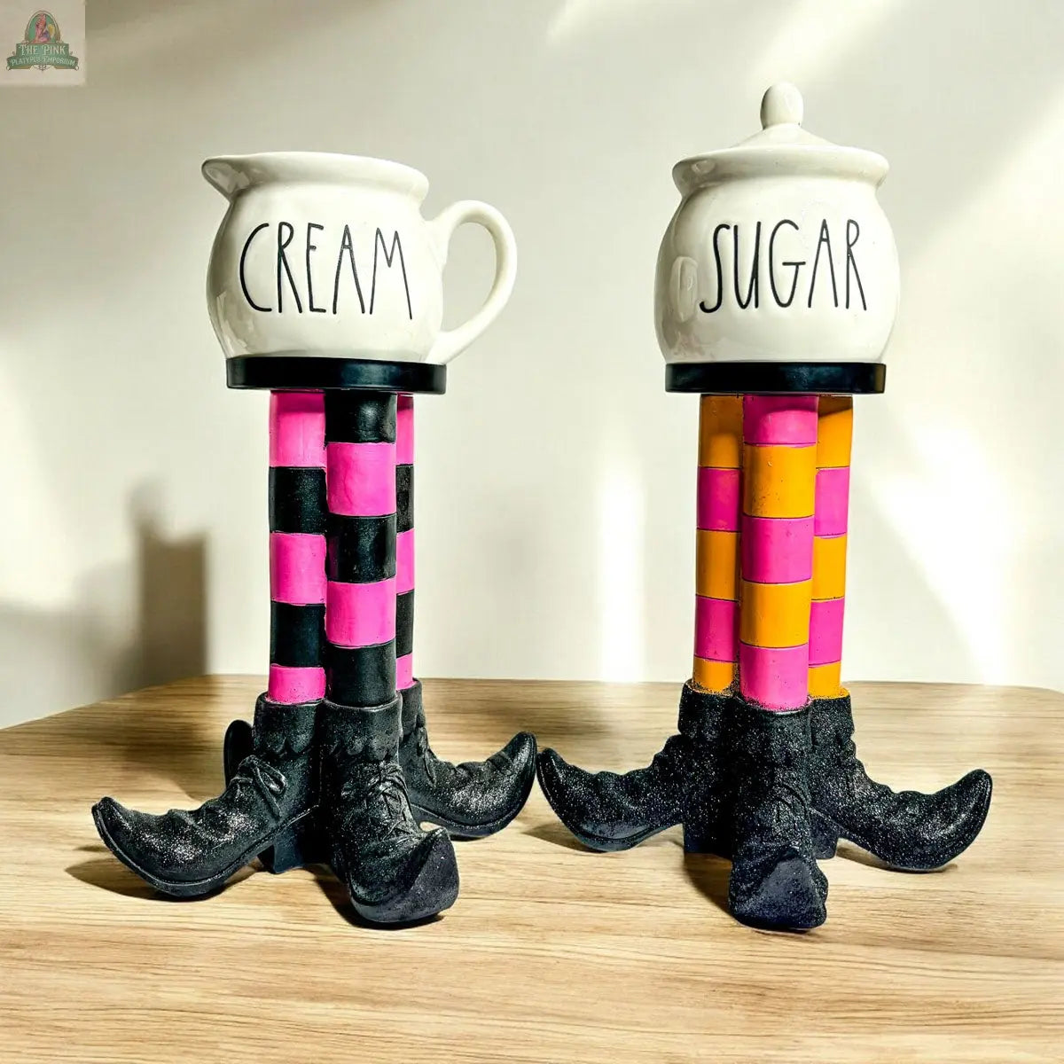 The Wicked Witch Candle Holder features two ceramic containers labeled CREAM and SUGAR atop tall bases shaped like striped witch legs and black shoes, creating a whimsical Halloween centerpiece for any wooden surface.
