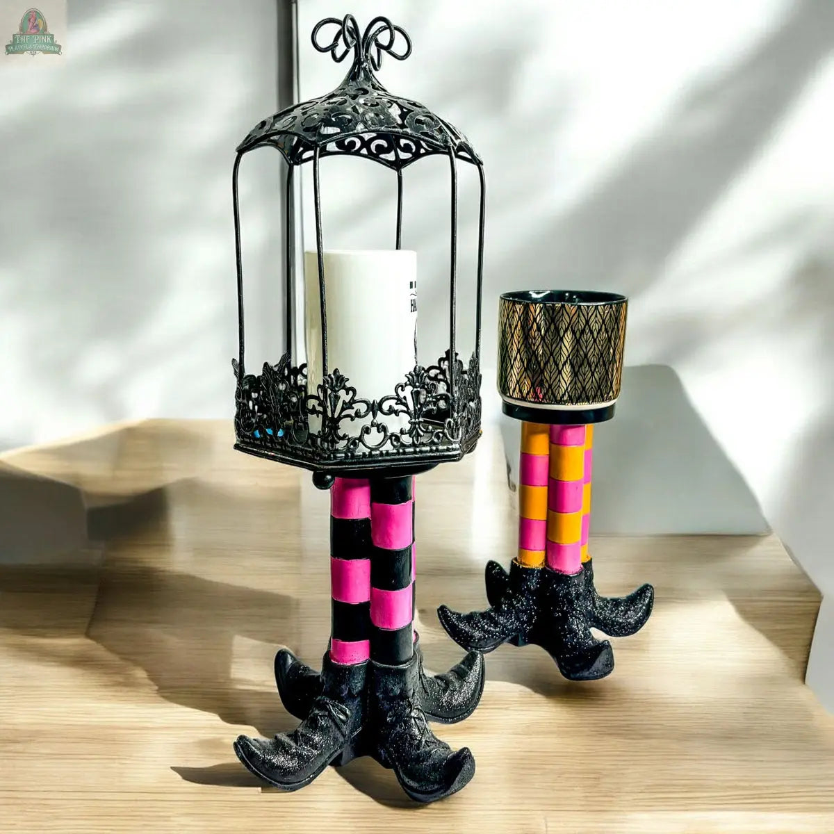 The Wicked Witch Candle Holder set features two decorative witch leg designs with striped bases, holding a white candle in a black ornate cage and a patterned cup. With pink, orange, and black accents, they're perfect for unique Halloween decor.