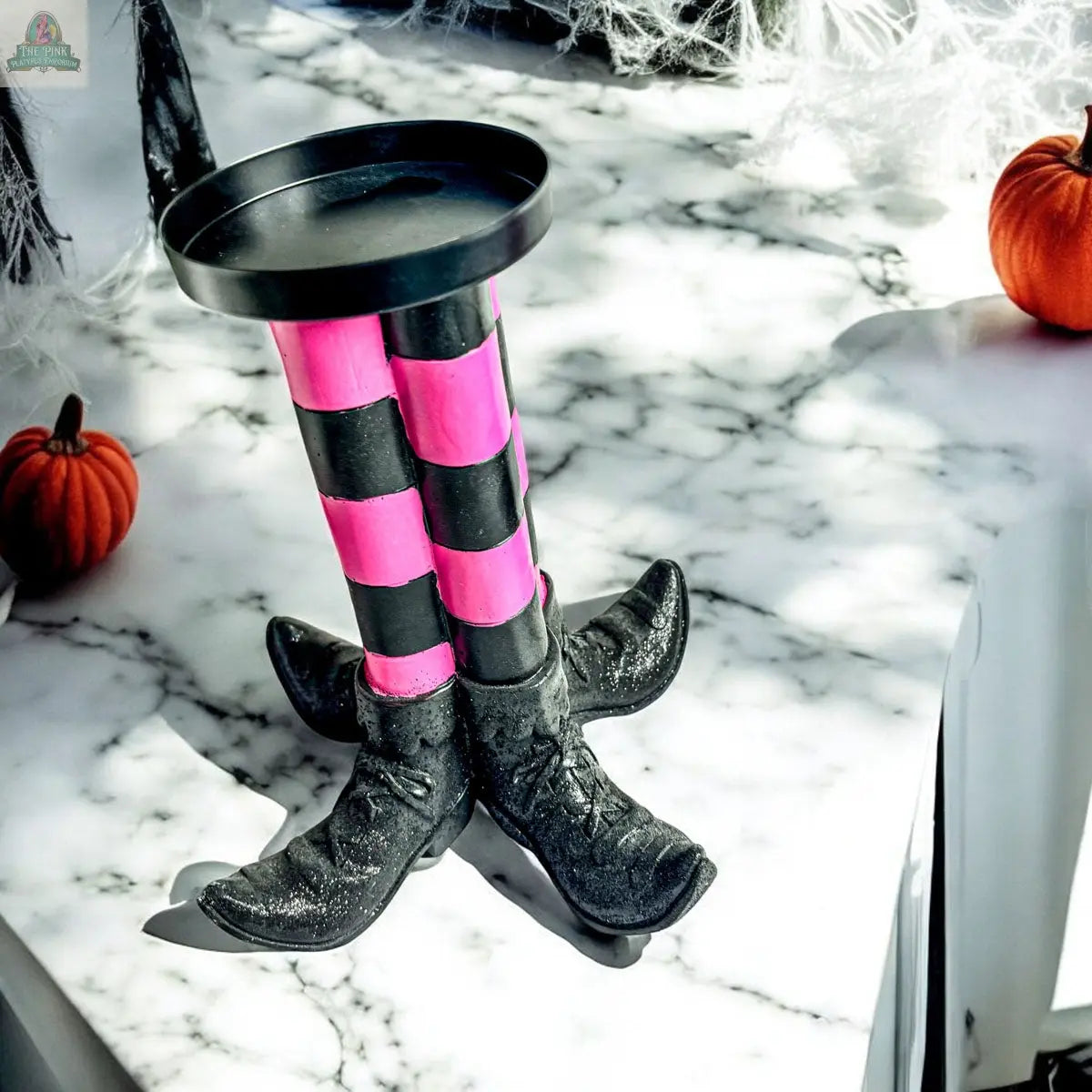 Add a touch of whimsy to your Halloween decor with the Wicked Witch Candle Holder, featuring black, pink, and purple striped legs in pointy shoes, displayed on a marble base with pumpkins and cobwebs—perfect as a unique centerpiece.
