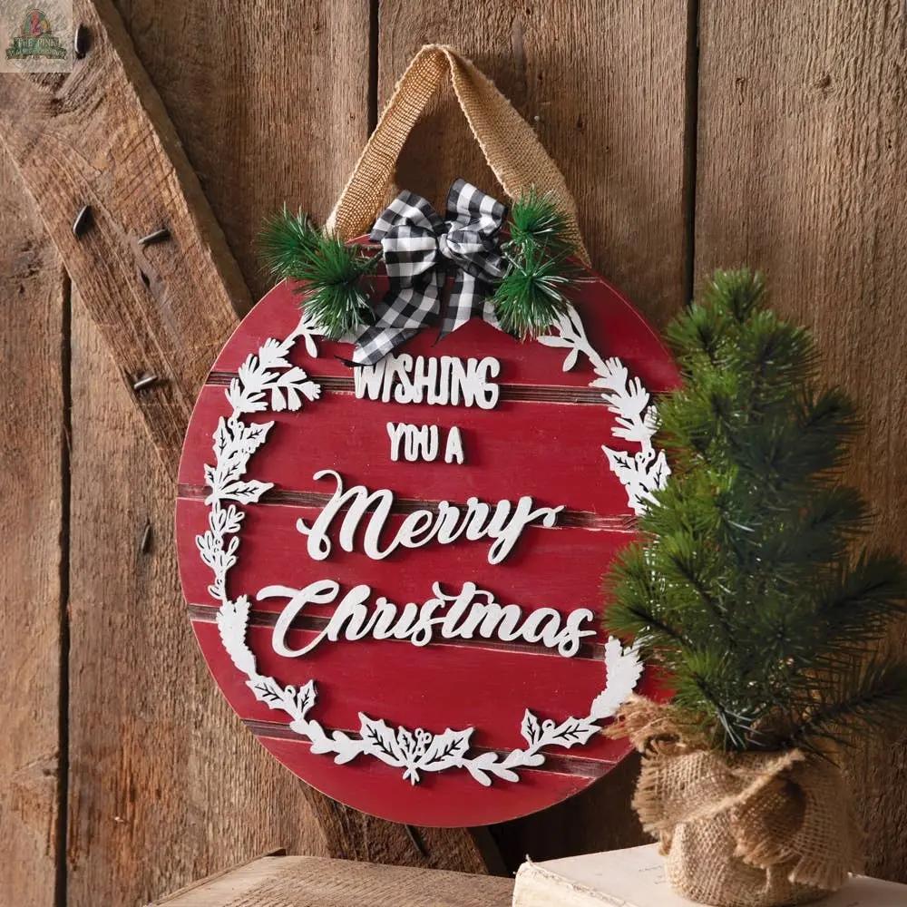 The Wishing You A Merry Christmas Ornament Sign features raised metal lettering, faux pine branches, and a plaid bow, hanging on rustic wood panels with a small burlap-wrapped pine tree nearby.