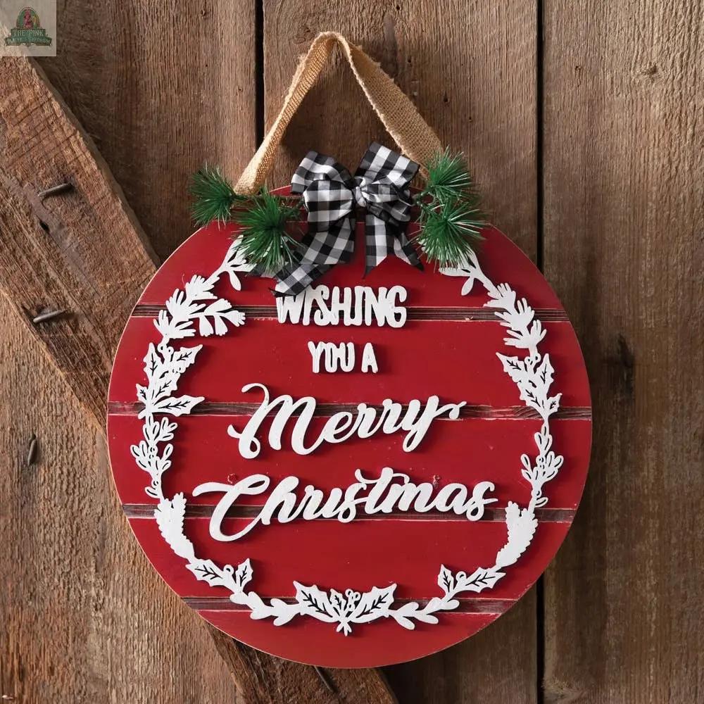 The Wishing You A Merry Christmas Ornament Sign is a round red metal sign with raised lettering and a wreath design. It hangs on a rope adorned with a plaid bow and pine accents, displayed against slatted wood panels.