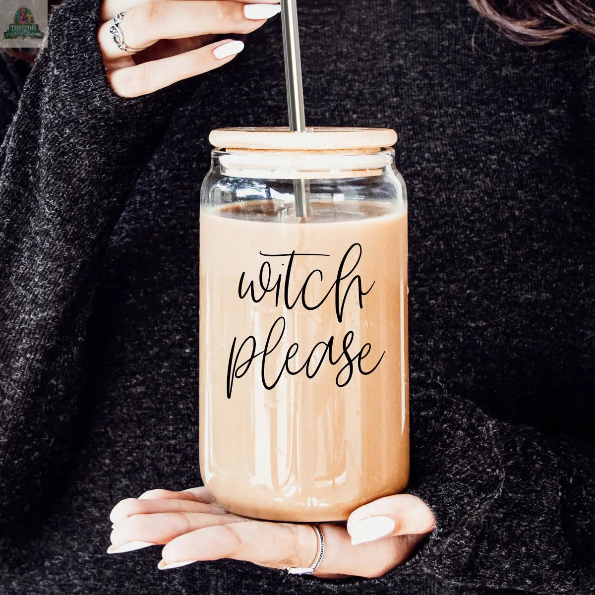 Someone in a dark sweater holds a 17oz Witch Please glass cup with a straw, filled with a light brown drink. The jar displays "witch please" in stylish black script.