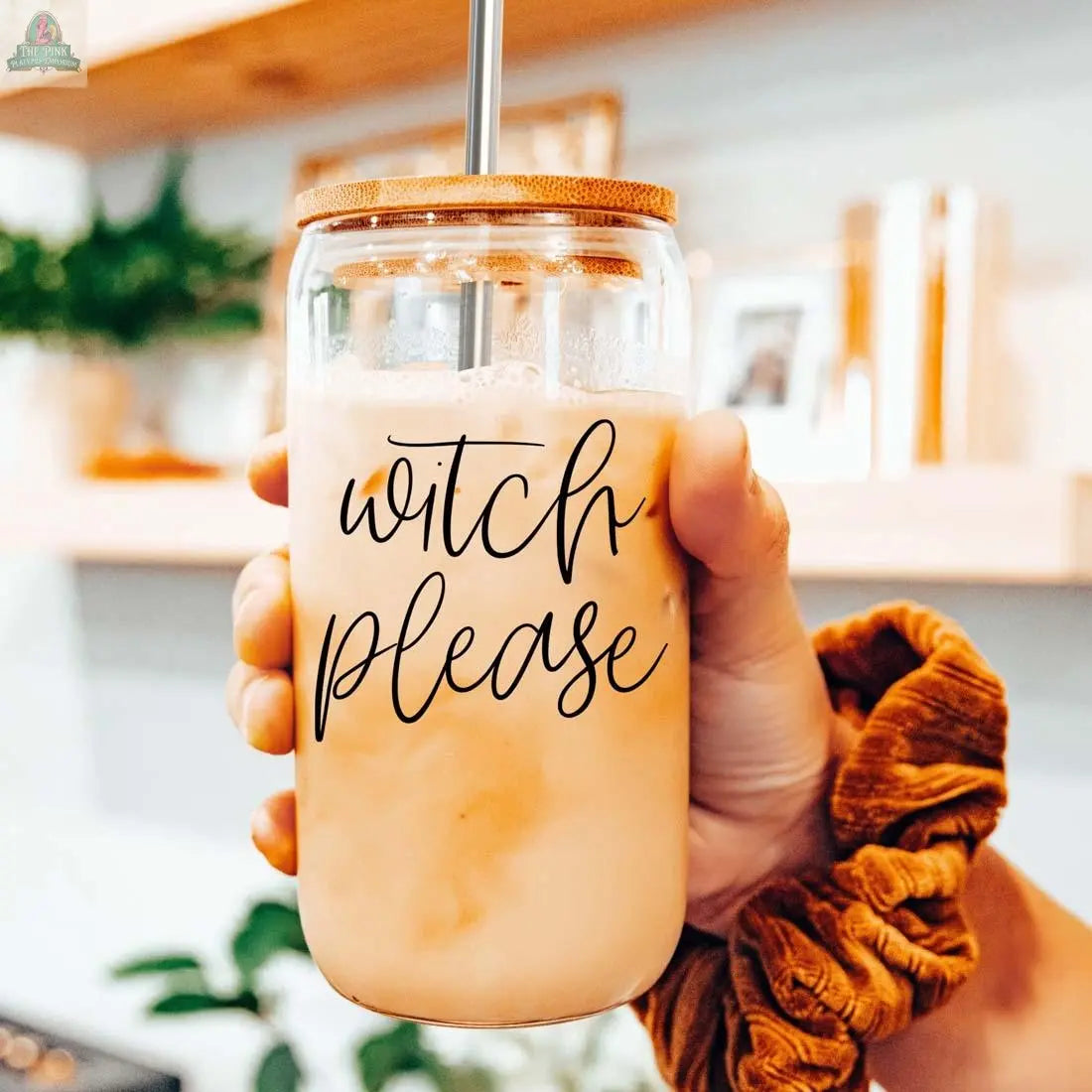A hand holds the Witch Please 17oz glass cup filled with iced coffee, featuring a bamboo lid and stainless steel straw. "Witch please" is displayed on the glass, with a brown scrunchie visible in a cozy kitchen setting.
