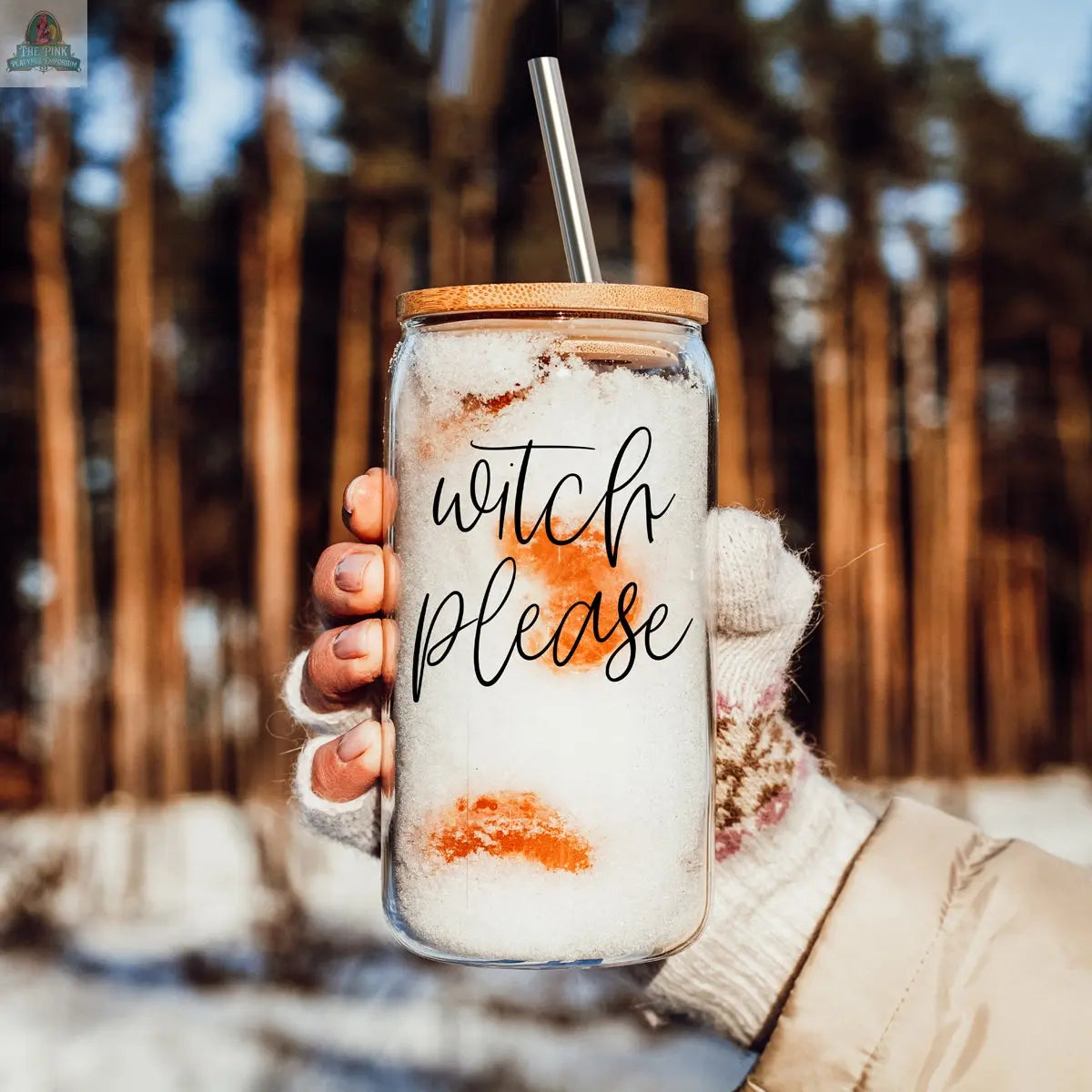 A hand in a patterned knit glove holds the Witch Please 17oz glass cup with a bamboo lid and stainless steel straw, filled with snow and orange slices. “Witch Please” is printed on the jar, with a snowy forest background.