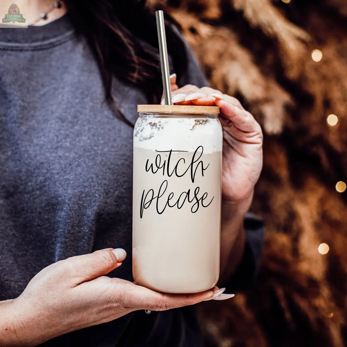 A hand holds the Witch Please 17oz glass jar, filled with a creamy drink topped with foam and a stainless steel straw. "Witch Please" is printed in black script on the jar, set against a softly blurred, warm brown background.