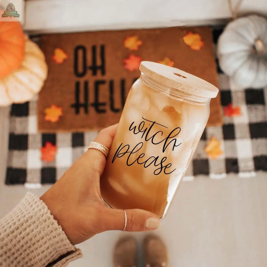 A hand holds the Witch Please 17oz glass cup with black script. In the background are pumpkins, autumn leaves, a checkered mat, and a doormat reading "OH HELLO.