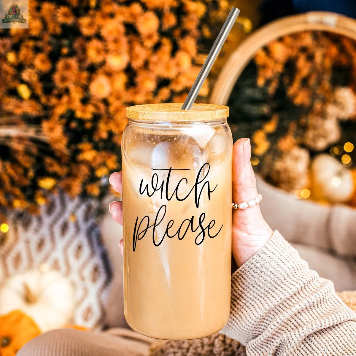 A hand holds the Witch Please 17oz glass cup filled with iced coffee and a stainless steel straw. Blurred autumn decor with orange flowers and pumpkins appears in the background.