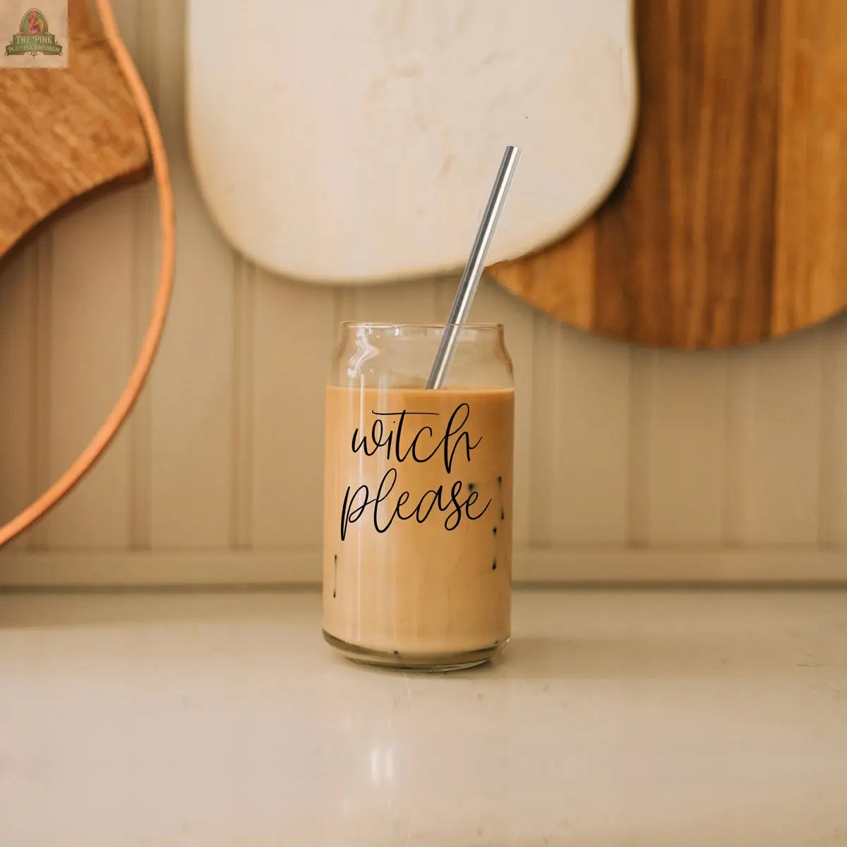 A Witch Please 17oz glass, ideal for glass drinking cup lovers, filled with iced coffee and a stainless steel straw, sits on a countertop. "Witch please" is written in cursive. Wooden cutting boards are blurred in the background.